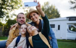Família jovem feliz com dois filhos tirando selfie com caravana ao fundo ao ar livre.